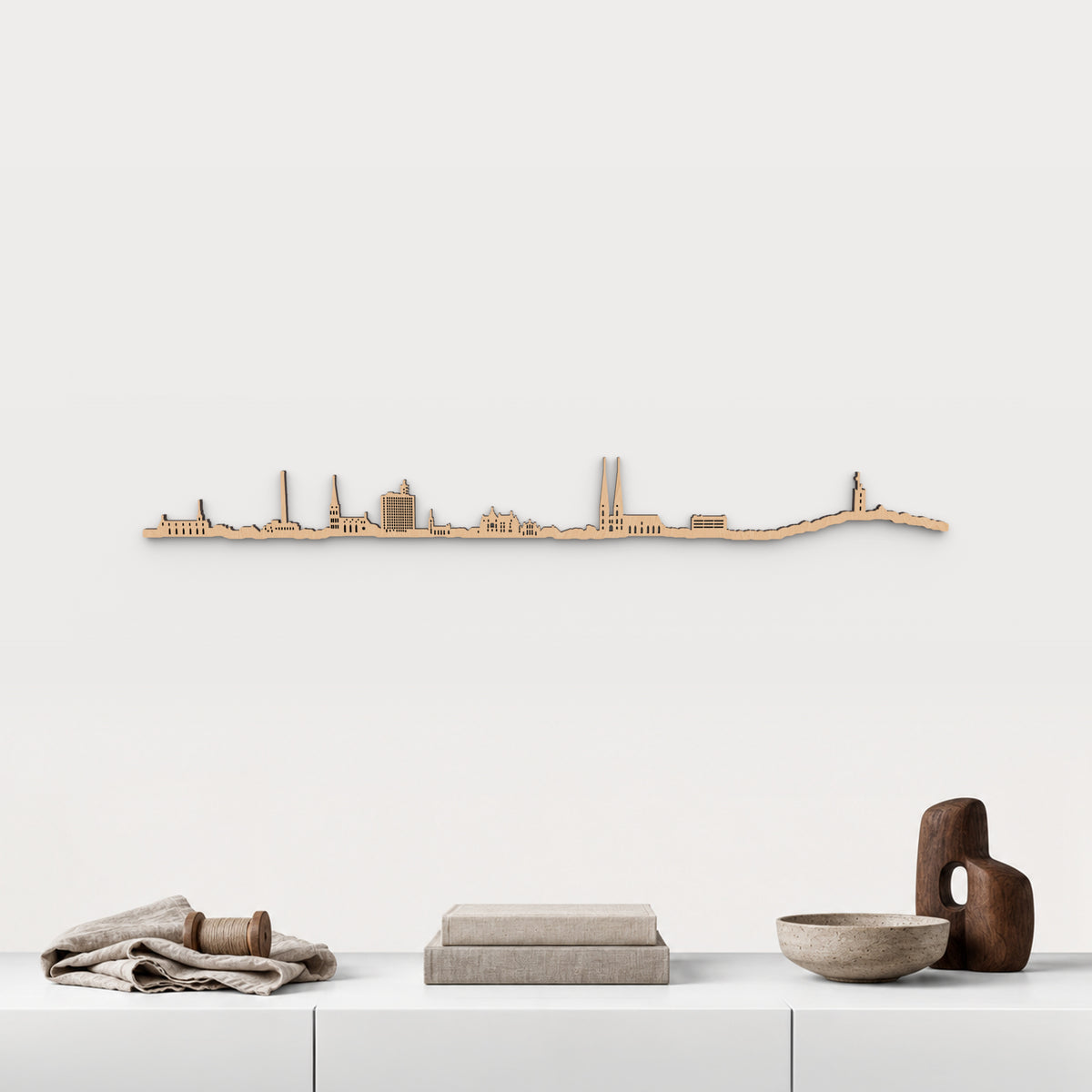 Skyline de Bielefeld en acabado marrón, presentado sobre el mueble blanco con lino, bobina de madera, libros y cerámica, en una decoración inspirada en la ciudad.