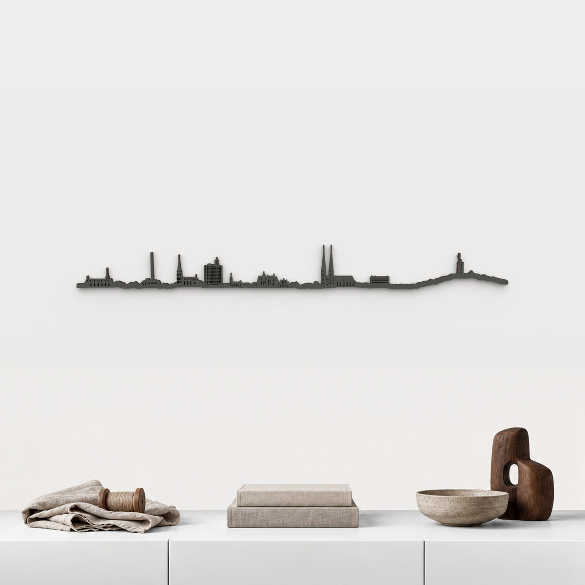 Skyline de Bielefeld en acabado negro, presentado sobre el mueble blanco con lino, bobina de madera, libros y cerámica, en una decoración inspirada en la ciudad.