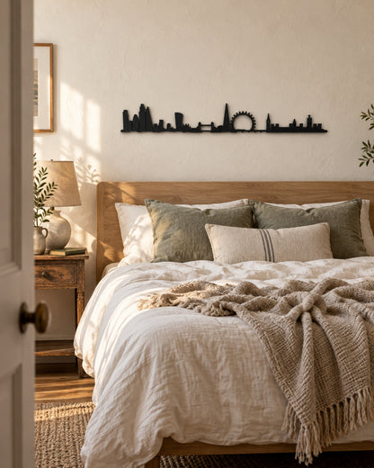 Skyline de Londres en madera negra sobre un cabecero de madera, en un dormitorio con ropa de cama clara y cojines verdes. Ambientación digital.