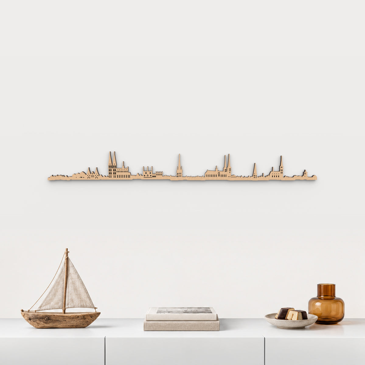 Skyline de Lübeck en acabado marrón, presentado sobre el mueble blanco con velero de madera, libros, mazapán y vidrio ámbar, en una decoración inspirada en la ciudad.