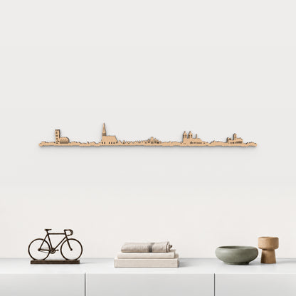 Skyline de Münster en acabado marrón, presentado sobre el mueble blanco con bicicleta decorativa, libros, lino y cerámica verde, en una decoración inspirada en la ciudad.