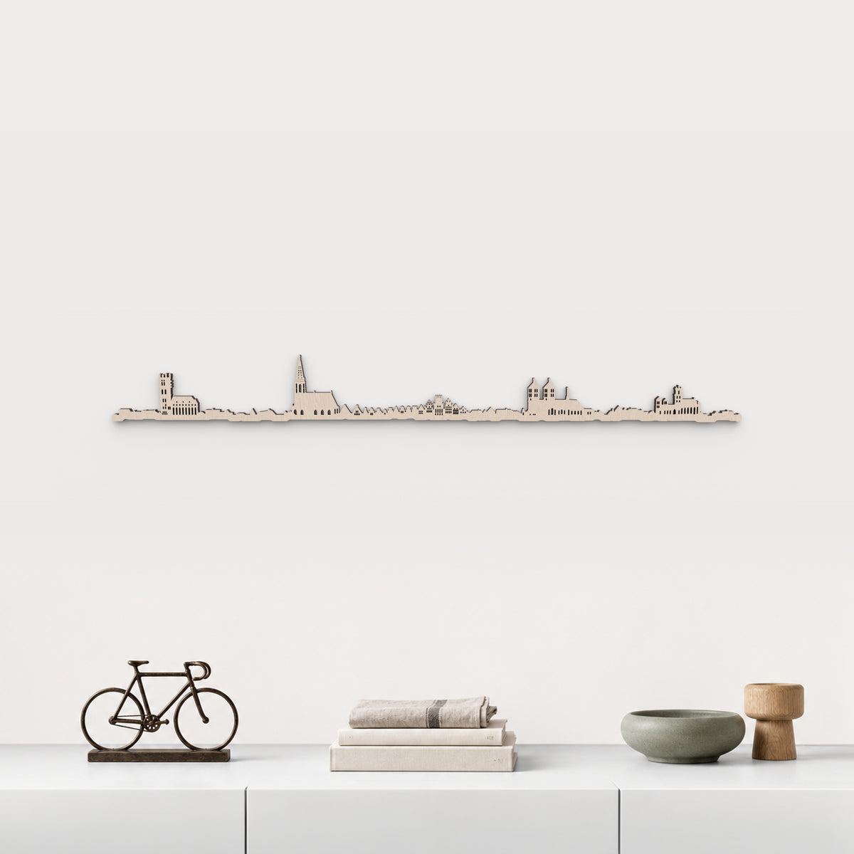 Skyline de Münster en acabado natural, presentado sobre el mueble blanco con bicicleta decorativa, libros, lino y cerámica verde, en una decoración inspirada en la ciudad.