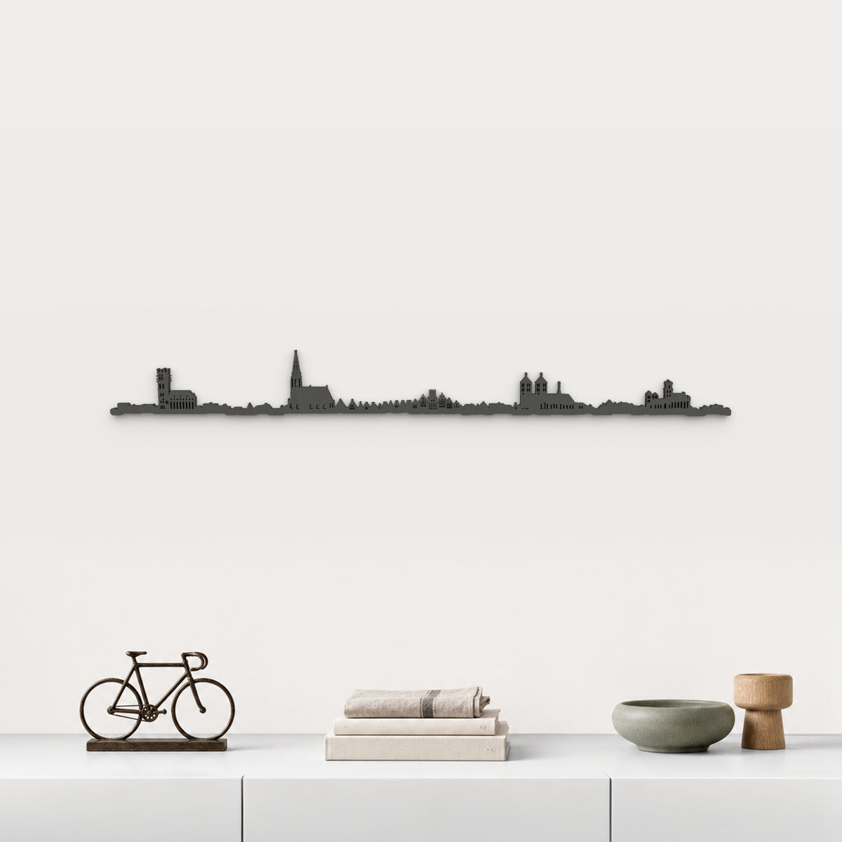 Skyline de Münster en acabado negro, presentado sobre el mueble blanco con bicicleta decorativa, libros, lino y cerámica verde, en una decoración inspirada en la ciudad.